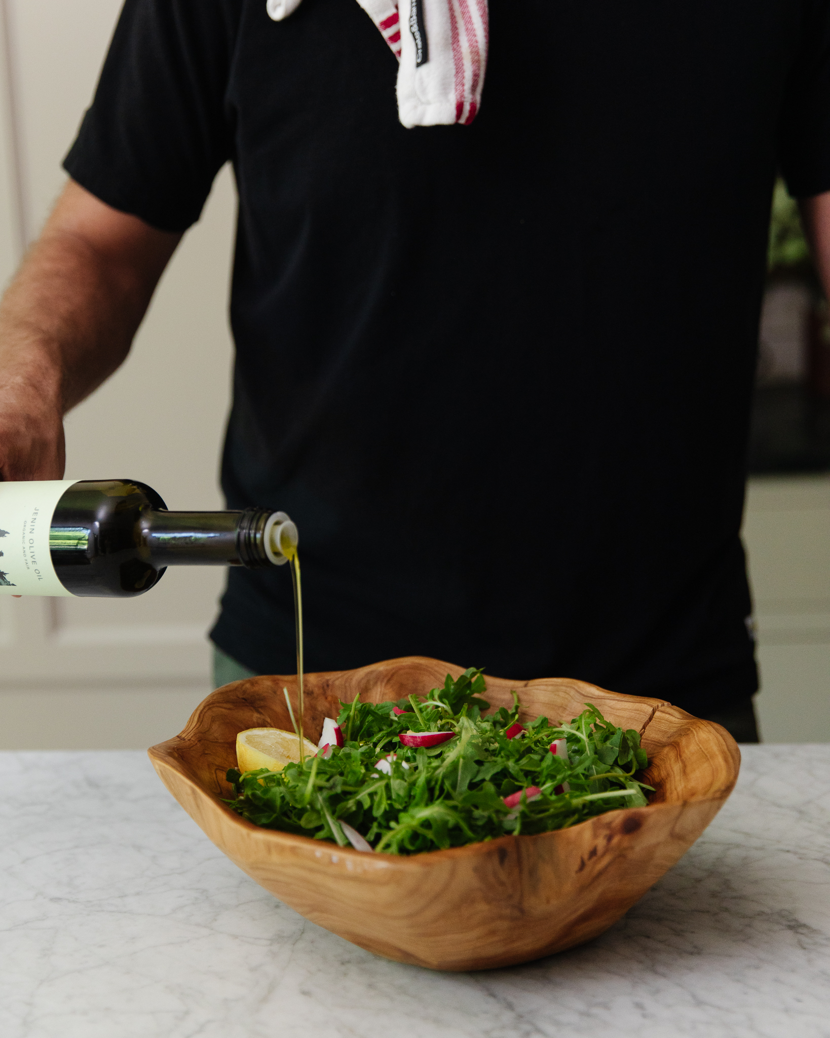 Chris Loves Julia | Chris pouring Canaan Jenin Olive Oil into a wooden bowl filled with arugula and radishes
