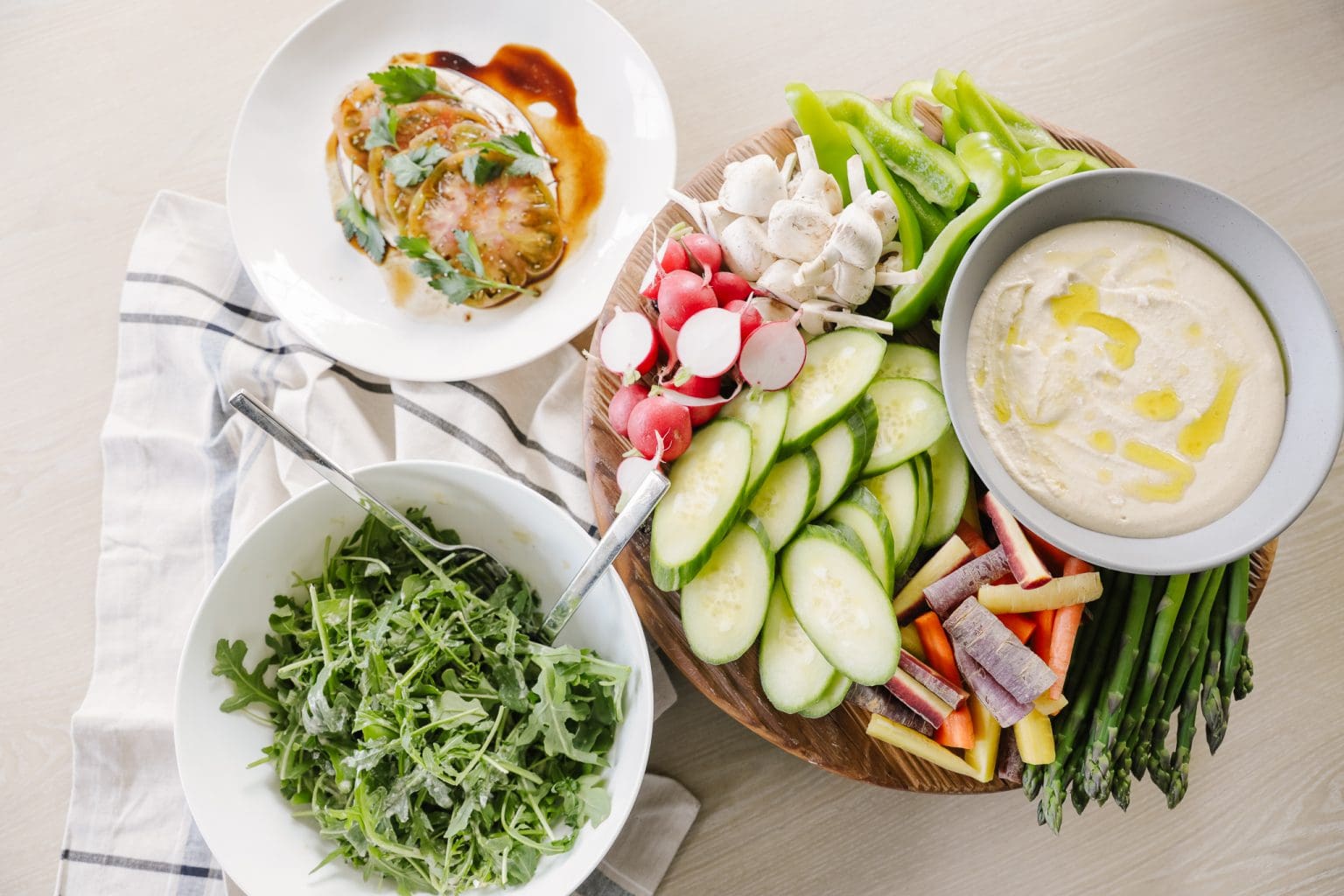 A bowl of hummus next to dippable veggies including asparagus, cucumbers, colorful carroets, radishes, and bell peppers.