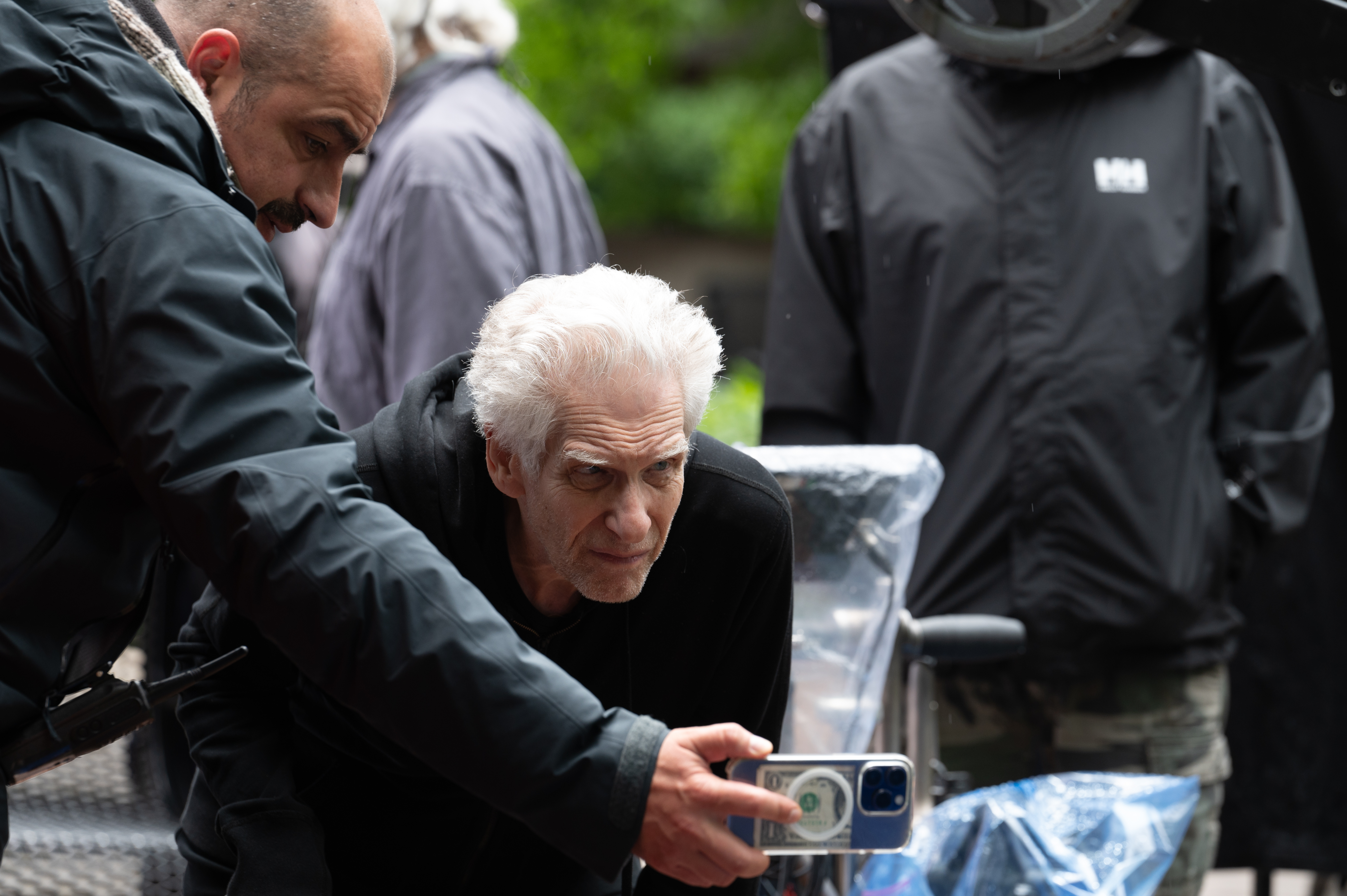 Film director David Cronenberg leaning forward to look at a camera screen held by an assistant