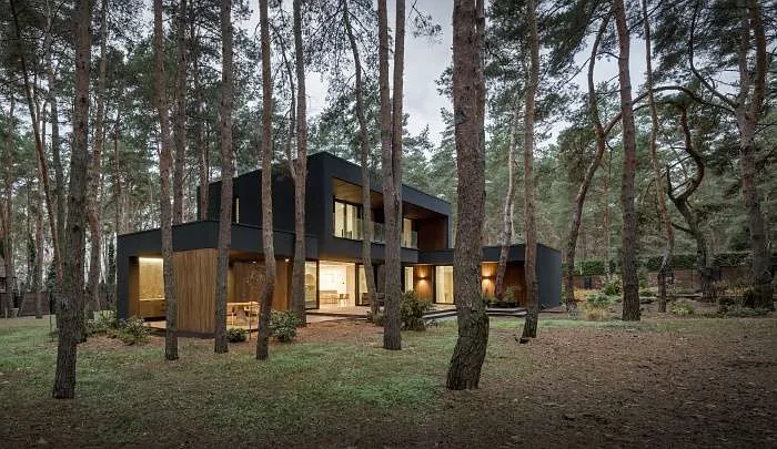 Night view of a black modern forest house glowing warmly among the trees.