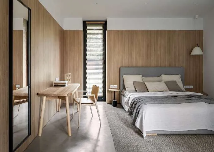 Bedroom with vertical wood panel walls, light textiles, and desk corner in a calm modern forest house interior.