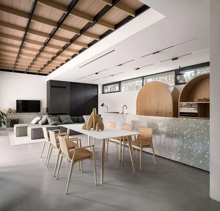 Open-plan kitchen and dining area with terrazzo island, arched wood cabinetry, and minimalist furniture in a modern forest house interior.