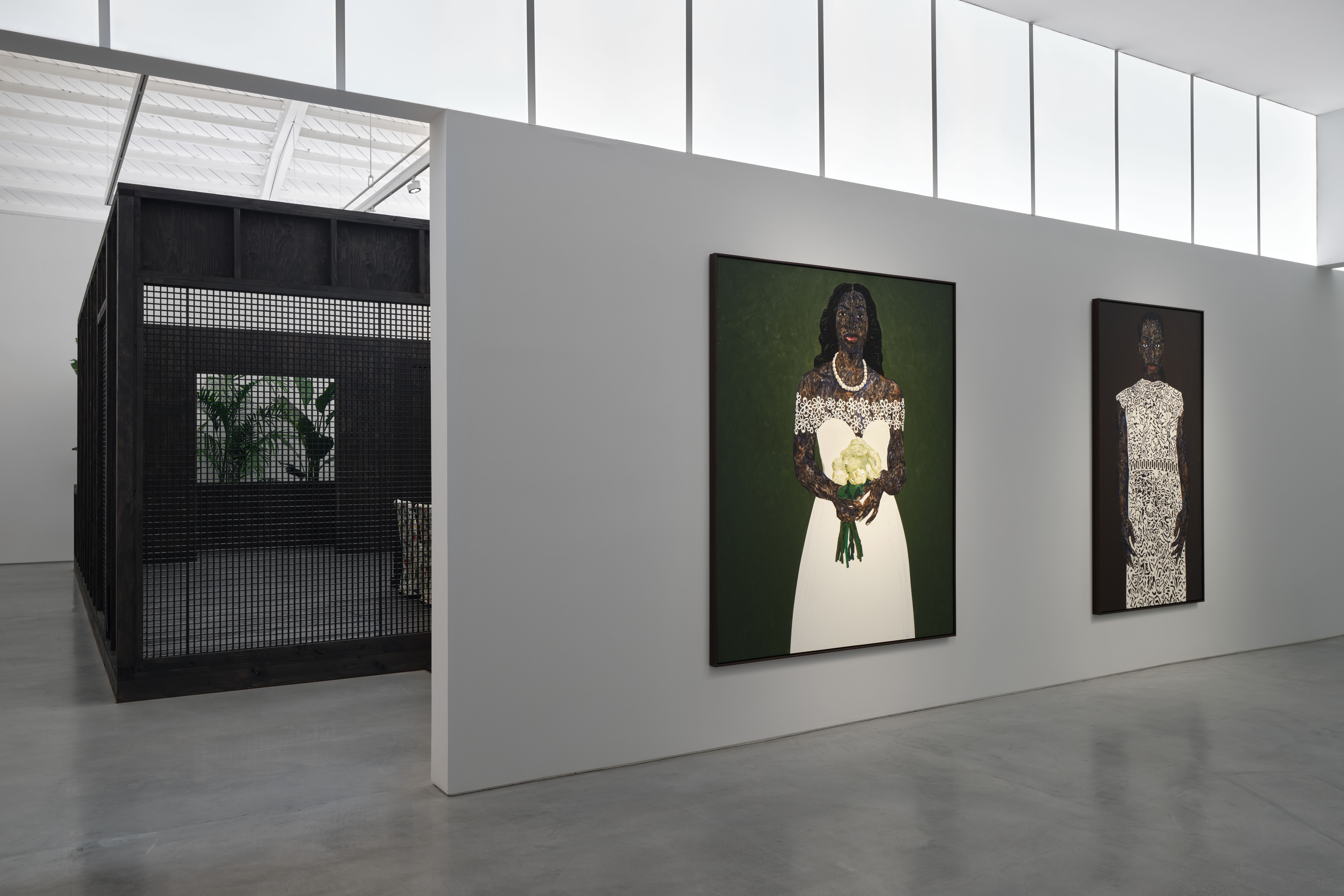 View of an art gallery installation with dark wood walls and multiple expressive portrait paintings. In the foreground, a large black metal structure with vertical bars frames additional paintings inside, while more portraits hang on the walls in the background.