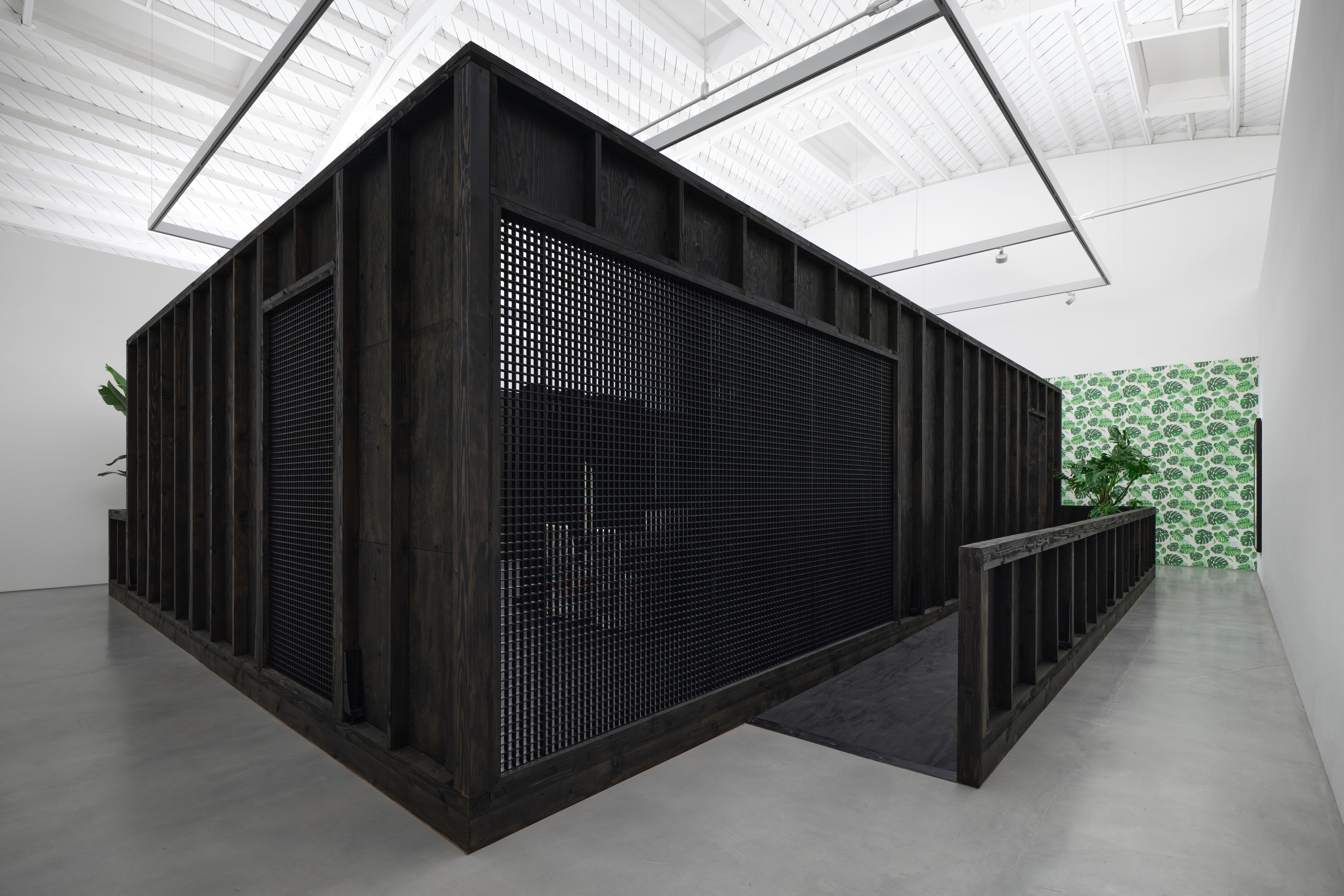 View of an art gallery installation with dark wood walls and multiple expressive portrait paintings. In the foreground, a large black metal structure with vertical bars frames additional paintings inside, while more portraits hang on the walls in the background.