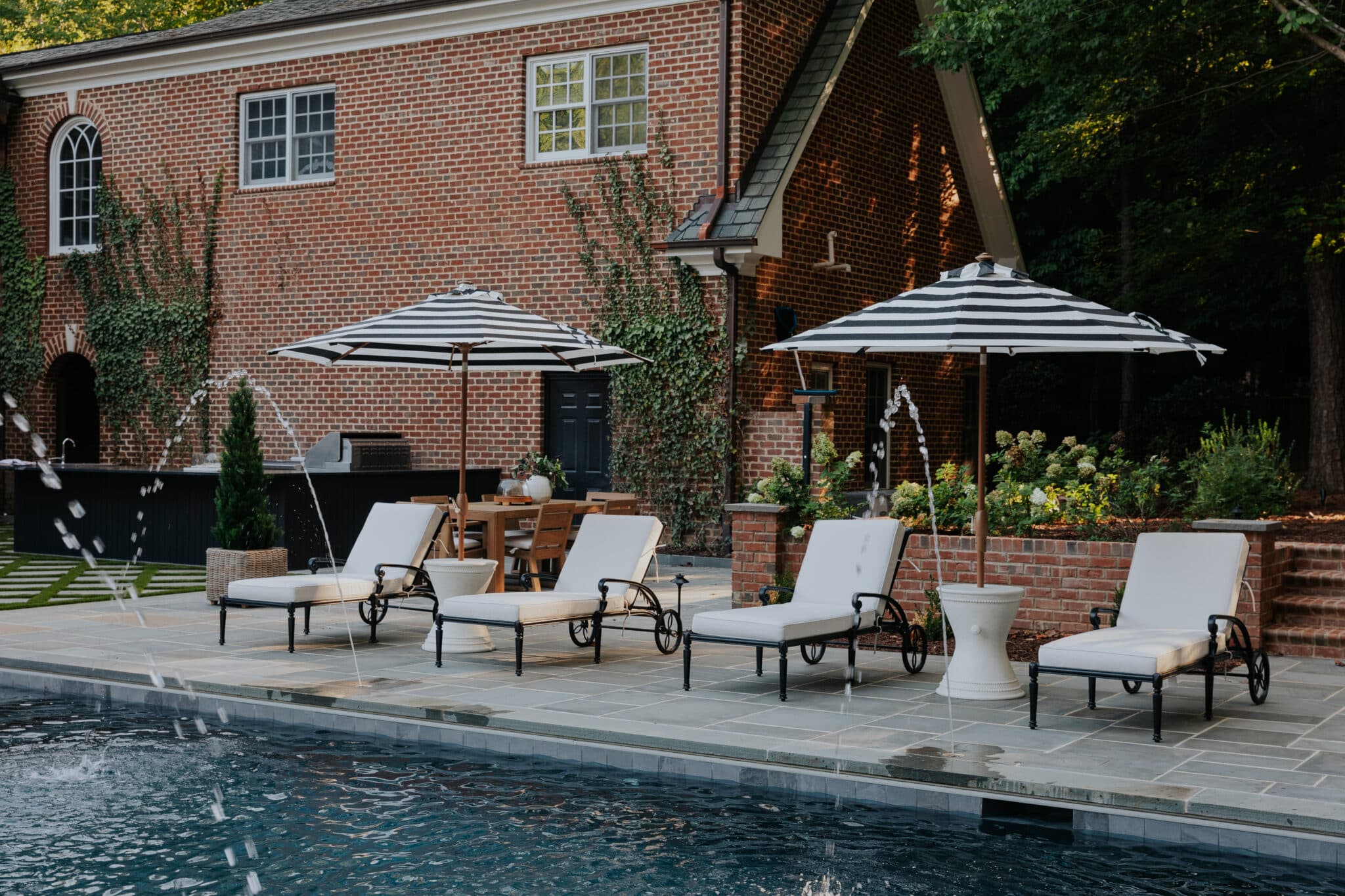 A dark-bottom luxury pool with arching water deck jets and four black metal chaise lounges with white cushions. Two black-and-white striped patio umbrellas are mounted on white side tables, with a brick home with ivy and an outdoor kitchen in the background.