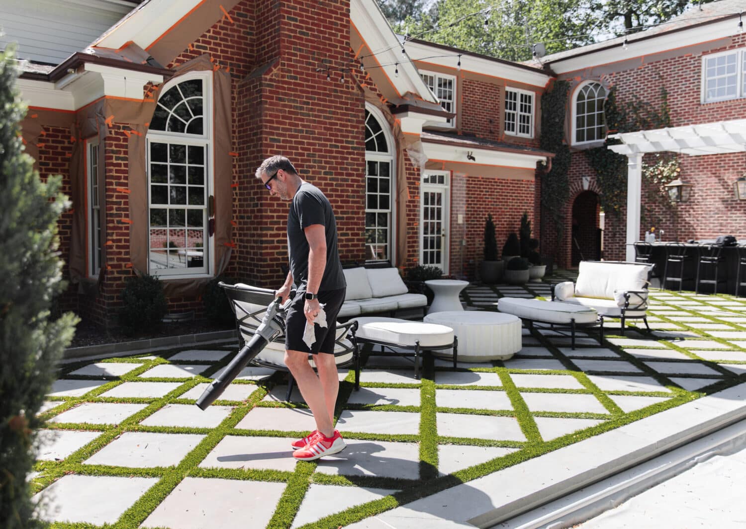 A person leaf-blowing a contemporary outdoor patio area with geometric grass and concrete tile design in front of a large brick house.