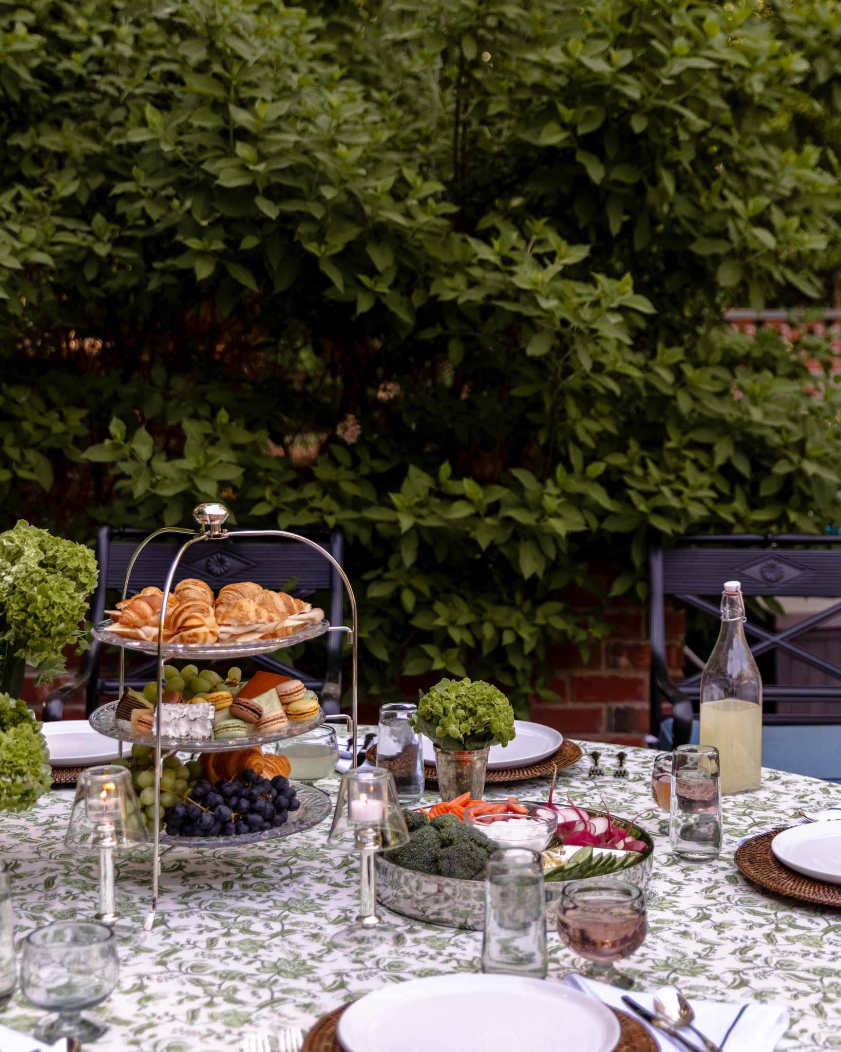 Outdoor dining table with a floral tablecloth, featuring a tiered serving tray, fresh fruit, and glassware.
