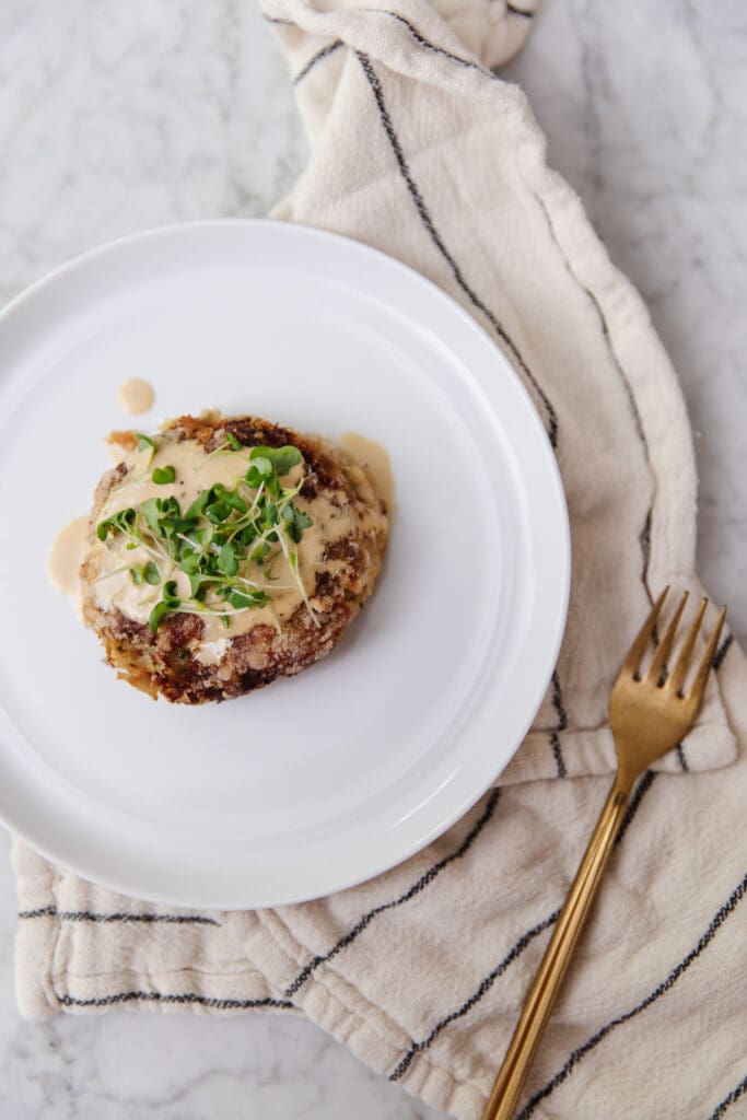 Delicious crab cake topped with microgreens on a white plate, served with a gold fork on a beige napkin.