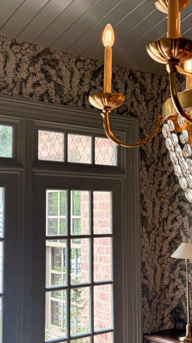Elegant chandelier with exposed bulbs in a cozy living space featuring floral wallpaper and a large window.
