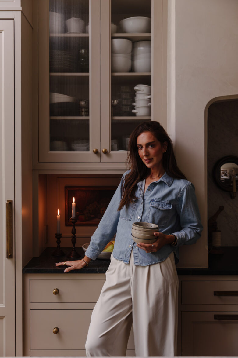 Julia Marcum standing in a cozy, well-organized kitchen holding a bowl, surrounded by open cabinets filled with white dishes and bowls, creating a warm and inviting atmosphere.